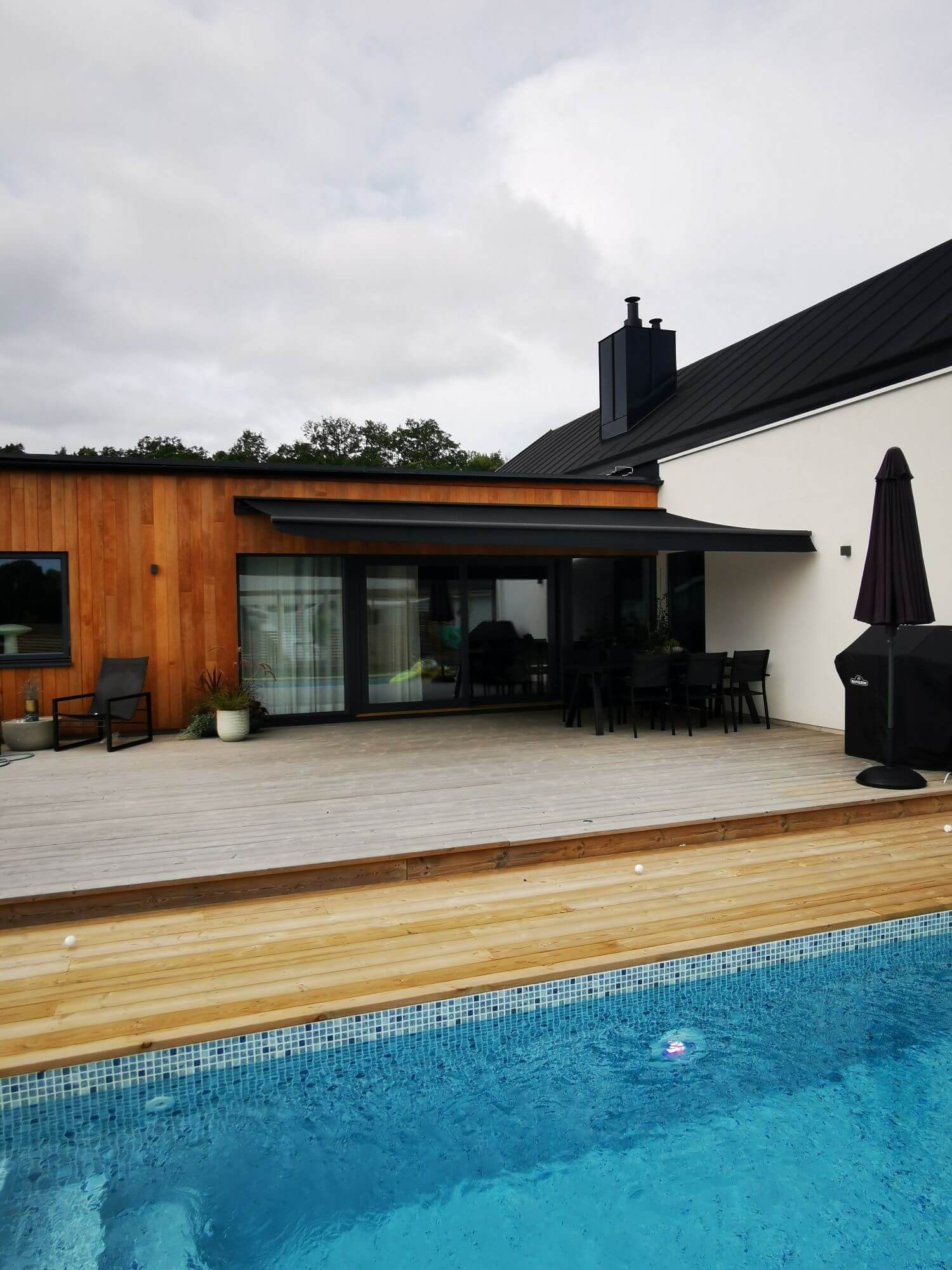 Jamaica inkl skyddshuv i Antracit - FSM71319 samt svart markisväv monterad över uteplatsen med tralldäck. Elektrisk styrning inklusive skaksensor som tillval för att markisen skall stängas när vinden börjar blåsa.