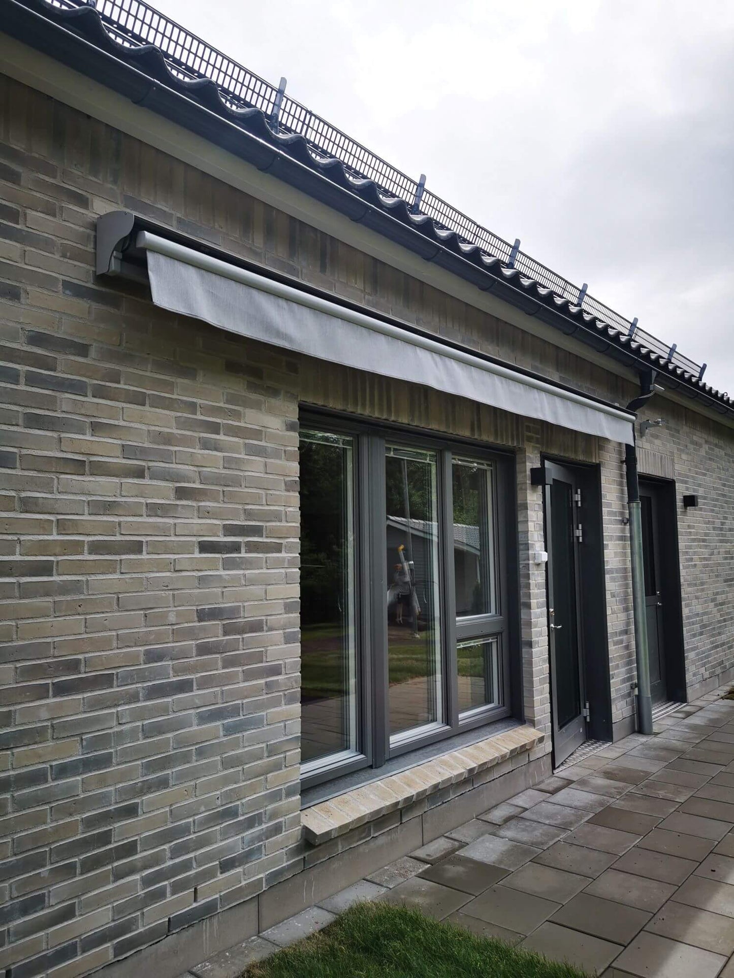 Jamaica inkl skyddshuv i grå kulör RAL9006 med ljusgrå markisväv monterad över uteplatsen. Elektrisk styrning.