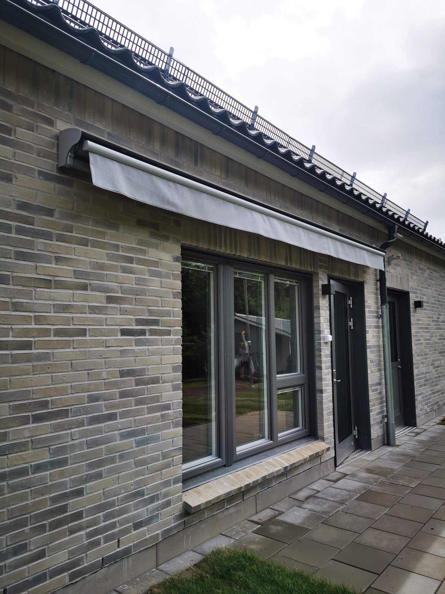 Jamaica inkl skyddshuv i grå kulör RAL9006 med ljusgrå markisväv monterad över uteplatsen. Elektrisk styrning.