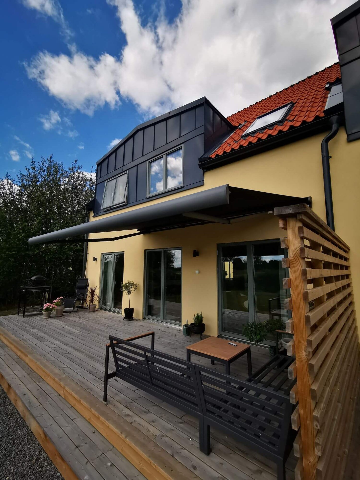 Antracit grå FSM71319 färgad markis med svart markisväv. Monterad på en gul putsad fasad vid uteplats med tralldäck och utemöbler. Premium Markis, modell Palladio. premium-markis.se