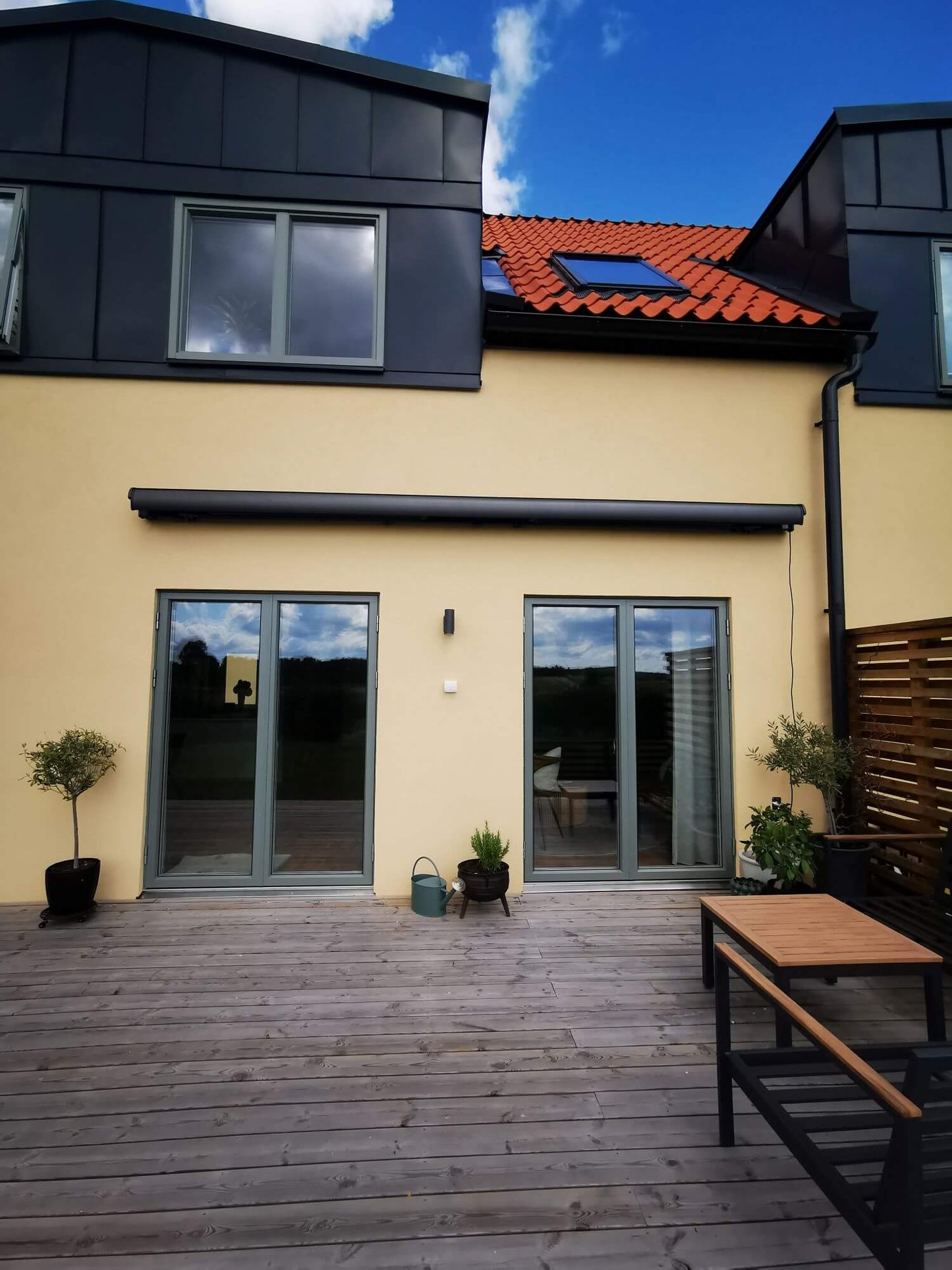 Antracit grå FSM71319 färgad markis med svart markisväv. Monterad på en gul putsad fasad vid uteplats med tralldäck och utemöbler. Premium Markis, modell Palladio. premium-markis.se
