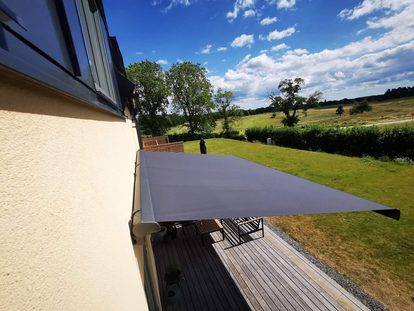 Antracit grå FSM71319 färgad markis med svart markisväv. Monterad på en gul putsad fasad vid uteplats med tralldäck och utemöbler. Premium Markis, modell Palladio. premium-markis.se