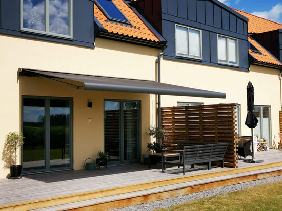 Antracit grå FSM71319 färgad markis med svart markisväv. Monterad på en gul putsad fasad vid uteplats med tralldäck och utemöbler. Premium Markis, modell Palladio. premium-markis.se