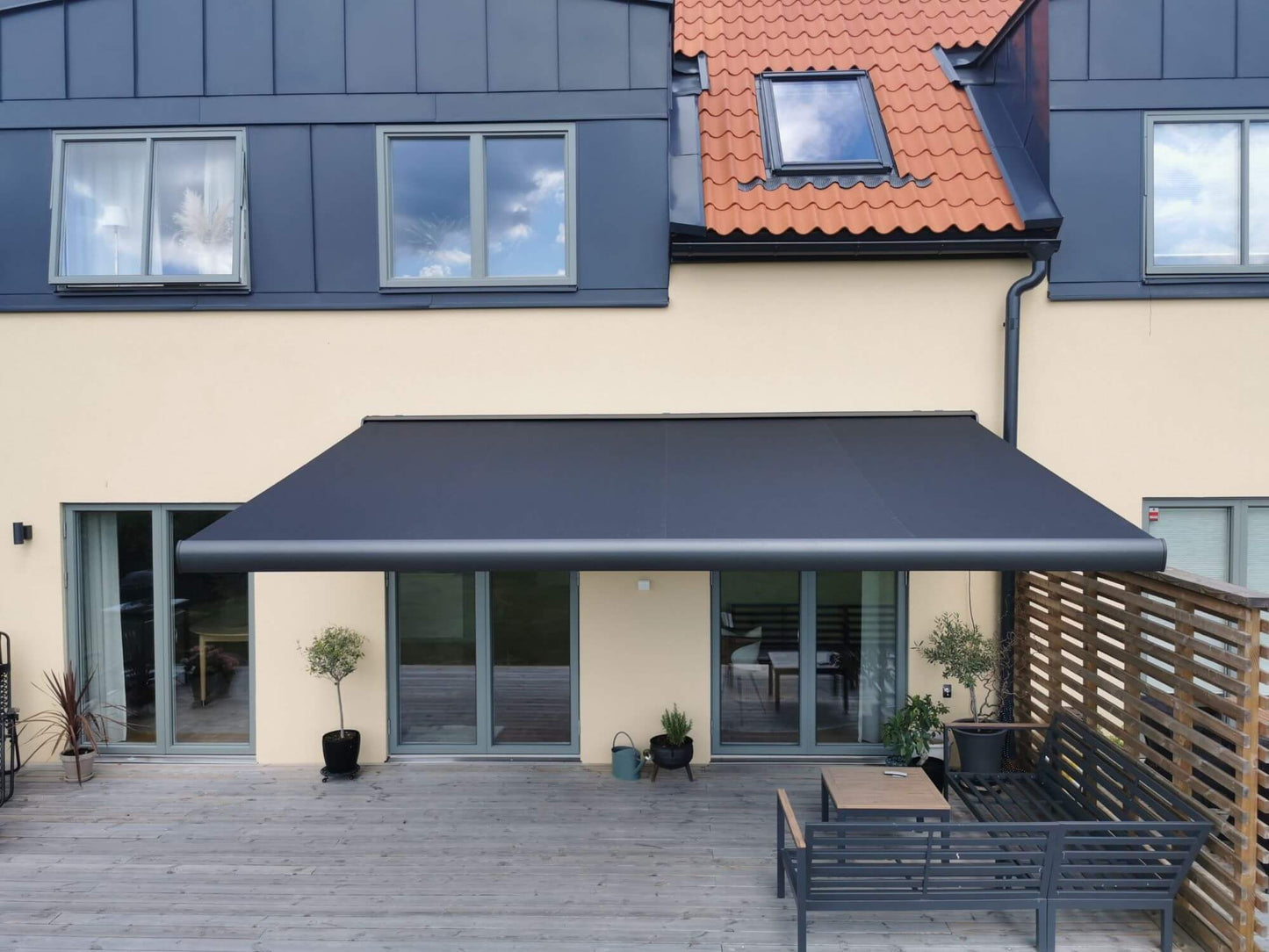 Antracit grå FSM71319 färgad markis med svart markisväv. Monterad på en gul putsad fasad vid uteplats med tralldäck och utemöbler. Premium Markis, modell Palladio. premium-markis.se